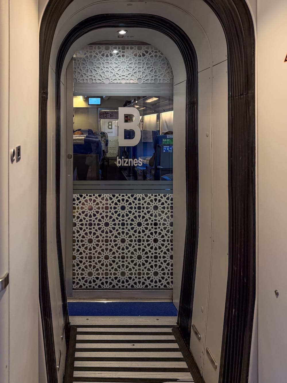 View through a rubber-sealed train gangway toward a glass door marked "B biznes" (business class), with geometric star-pattern frosted glass on the lower panel and blue seats visible in the carriage beyond.