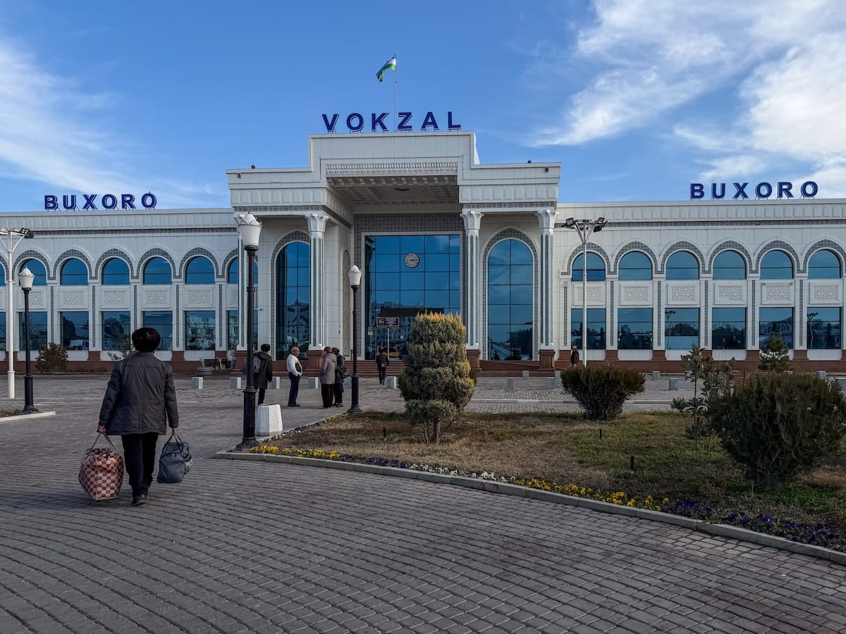 Exterior of Bukhara railway station in Uzbekistan, a white building with arched blue-glass windows and "VOKZAL" and "BUXORO" signs on the roofline, seen from a paved plaza where a traveler carries two bags toward the entrance.