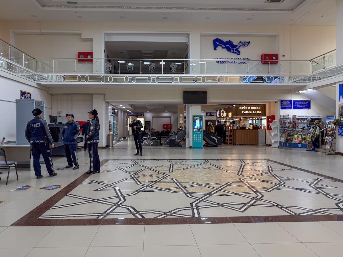 Spacious railway station hall with a geometric star-patterned marble floor, a mezzanine balcony carrying a blue Silk Road map and the words "Discover Silk Road with Uzbekistan Railways," a security checkpoint with uniformed officers on the left, and a "Coffee to Go" kiosk and souvenir stand at the back.