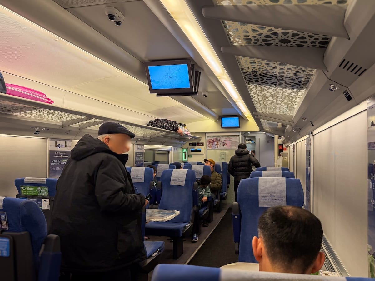 Interior of a business-class train carriage with blue seats and headrest covers reading "Uzbekistan Railways," ceiling-mounted screens showing route maps, geometric-pattern panels above the luggage racks, and passengers seated and standing along the aisle.