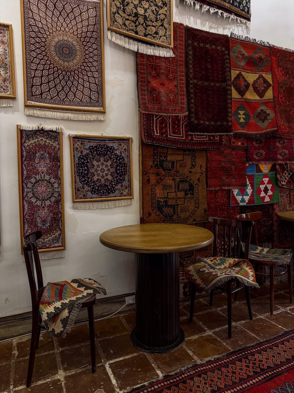Magic House coffee shop in Bukhara
table and chairs. with carpets on a wall behind it in uzbekistan