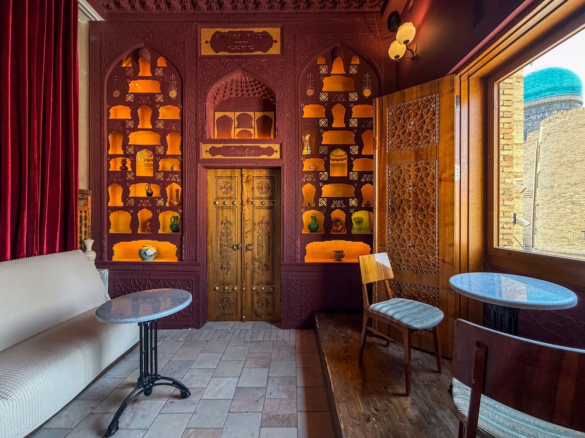 Pavillon Kalan, one of the most famous cafes in Bukhara the interior of a cafe in uzbekistan that has a couch on one side and tables by windows overlooking a square on the other side