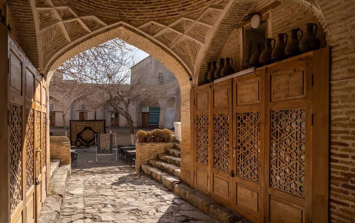 Brick archway in Bukhara’s old town leading to a stone-paved courtyard, with carved wooden lattice doors, clay jugs on top, and carpets hung in the background
