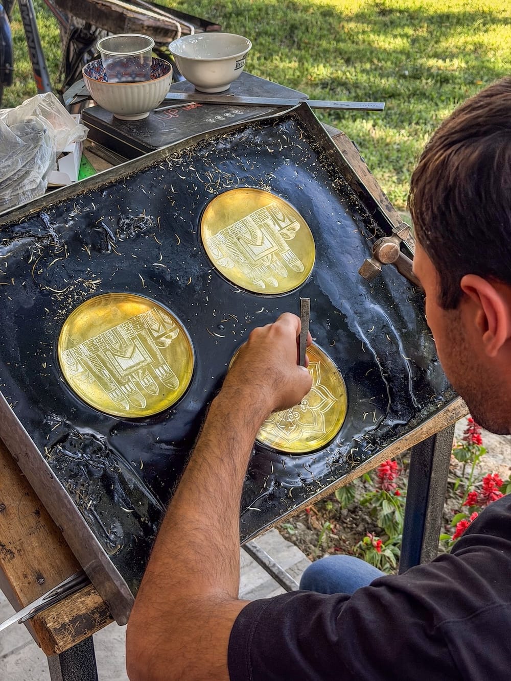 Metal engraver hand-chiseling a pattern into brass plates set in a black pitch tray at a metalwork workshop in Bukhara