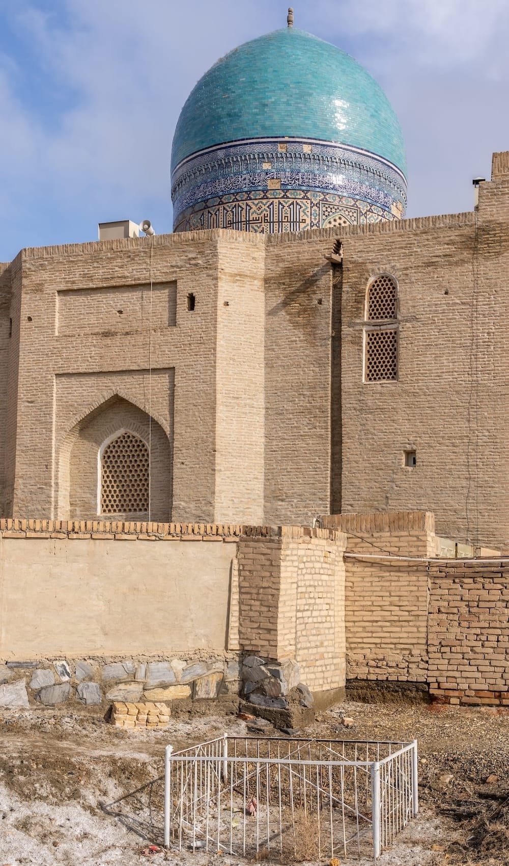 Turquoise tiled dome rising above the brick walls of a historic building in Bukhara, seen from a quiet, dusty lot on a winter day