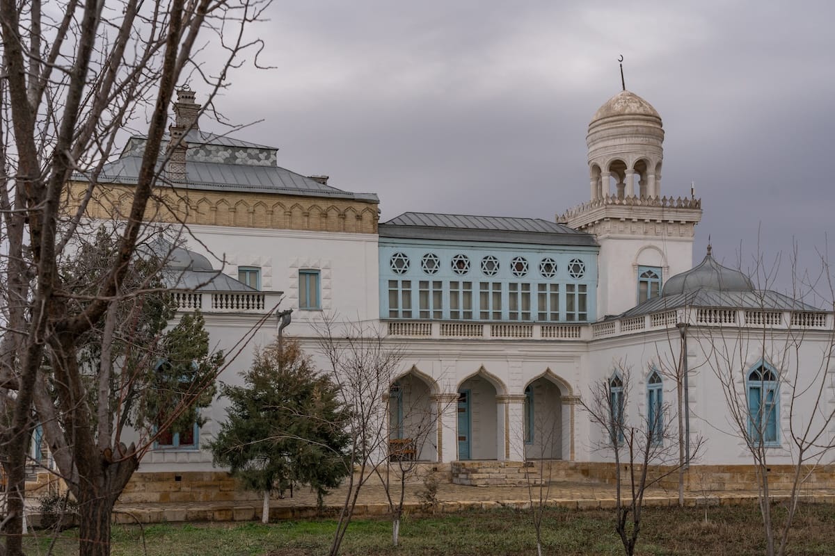 White palace with blue windows, arched entrance, and a domed tower surrounded by bare trees under a gray winter sky near Bukhara