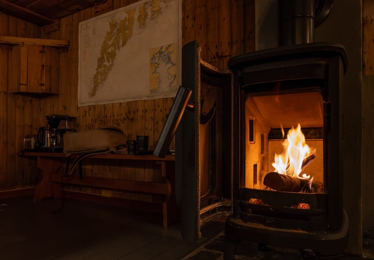 Wood-burning stove with a lit fire inside a cozy wood-paneled rorbu at Sakrisøy Rorbuer, Lofoten, with a map of the islands on the wall
