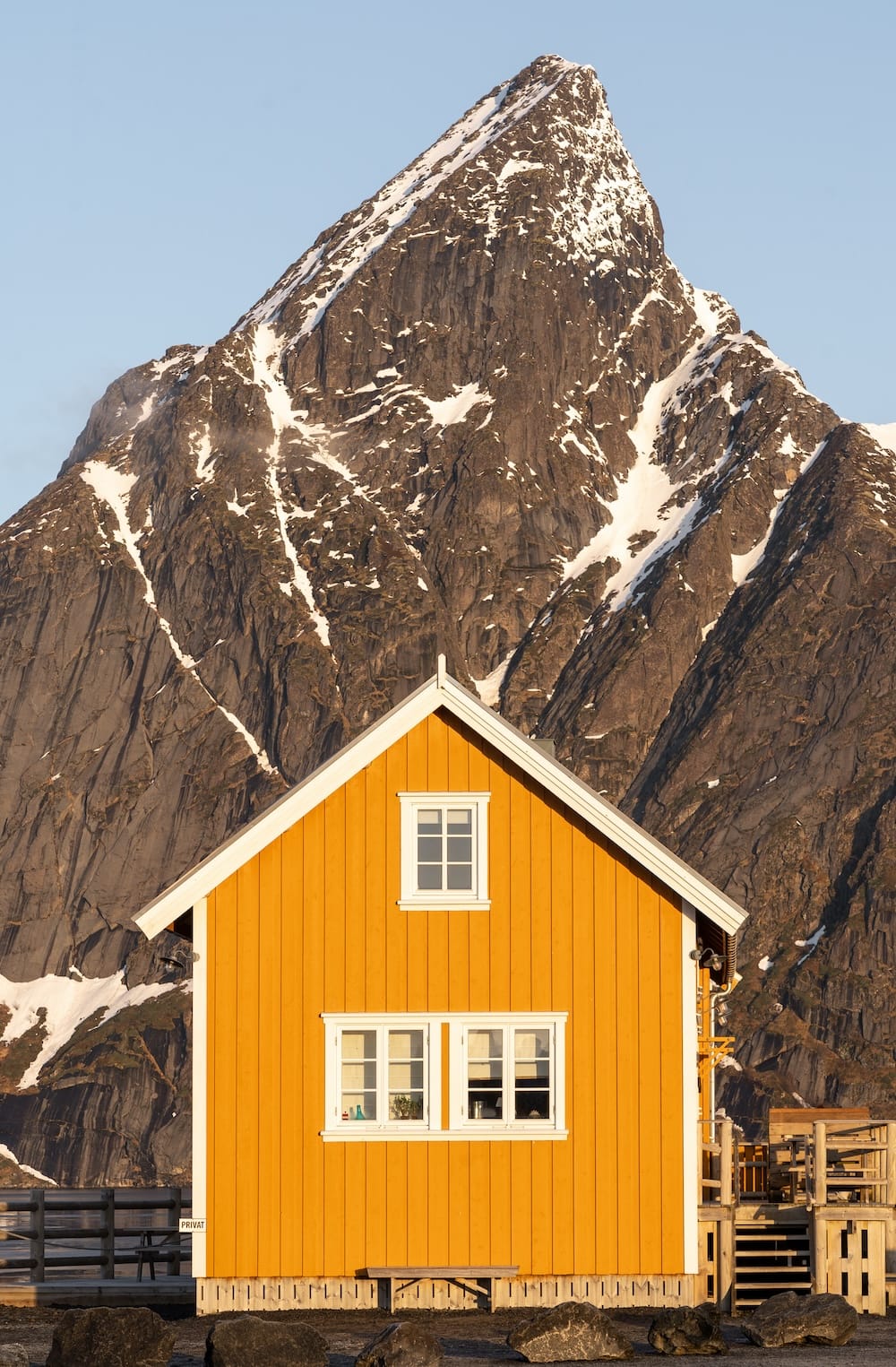 Yellow rorbu cabin in golden light with a steep, snow-streaked mountain peak rising behind it, likely at Sakrisøy, Lofoten