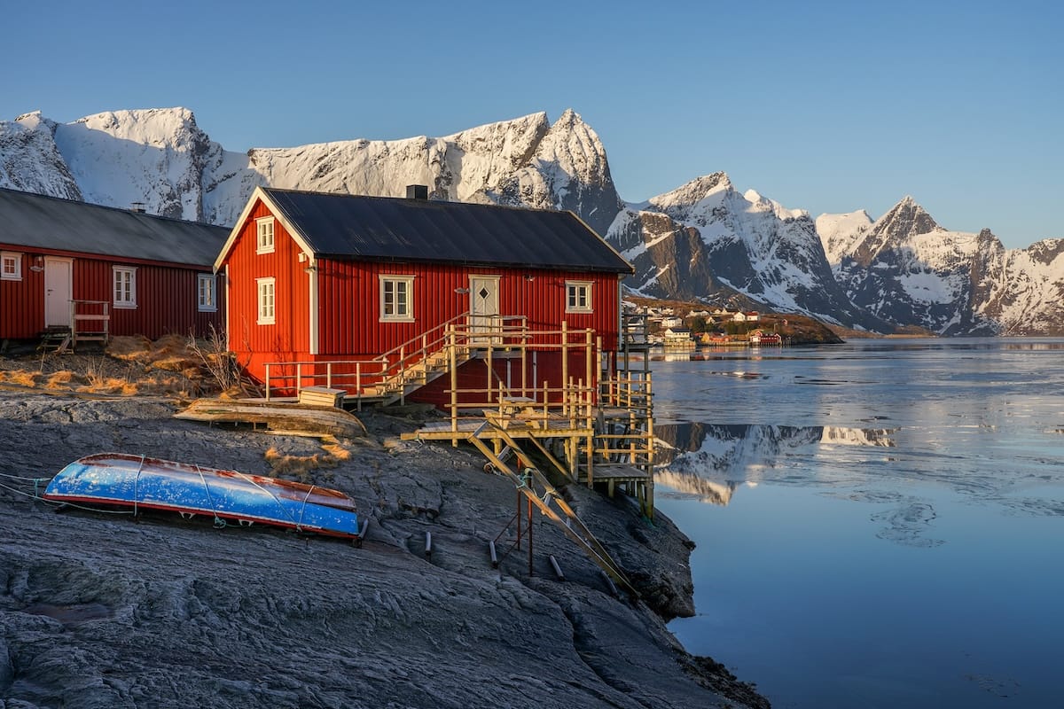 Red rorbu cabin on stilts beside a blue upturned boat on the rocky shore of a calm fjord with snow-capped Lofoten mountains behind