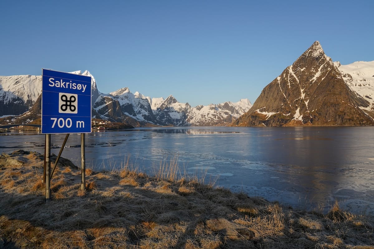 Blue road sign reading Sakrisøy 700 m beside a calm, partly frozen fjord with snow-capped Lofoten mountains in the background