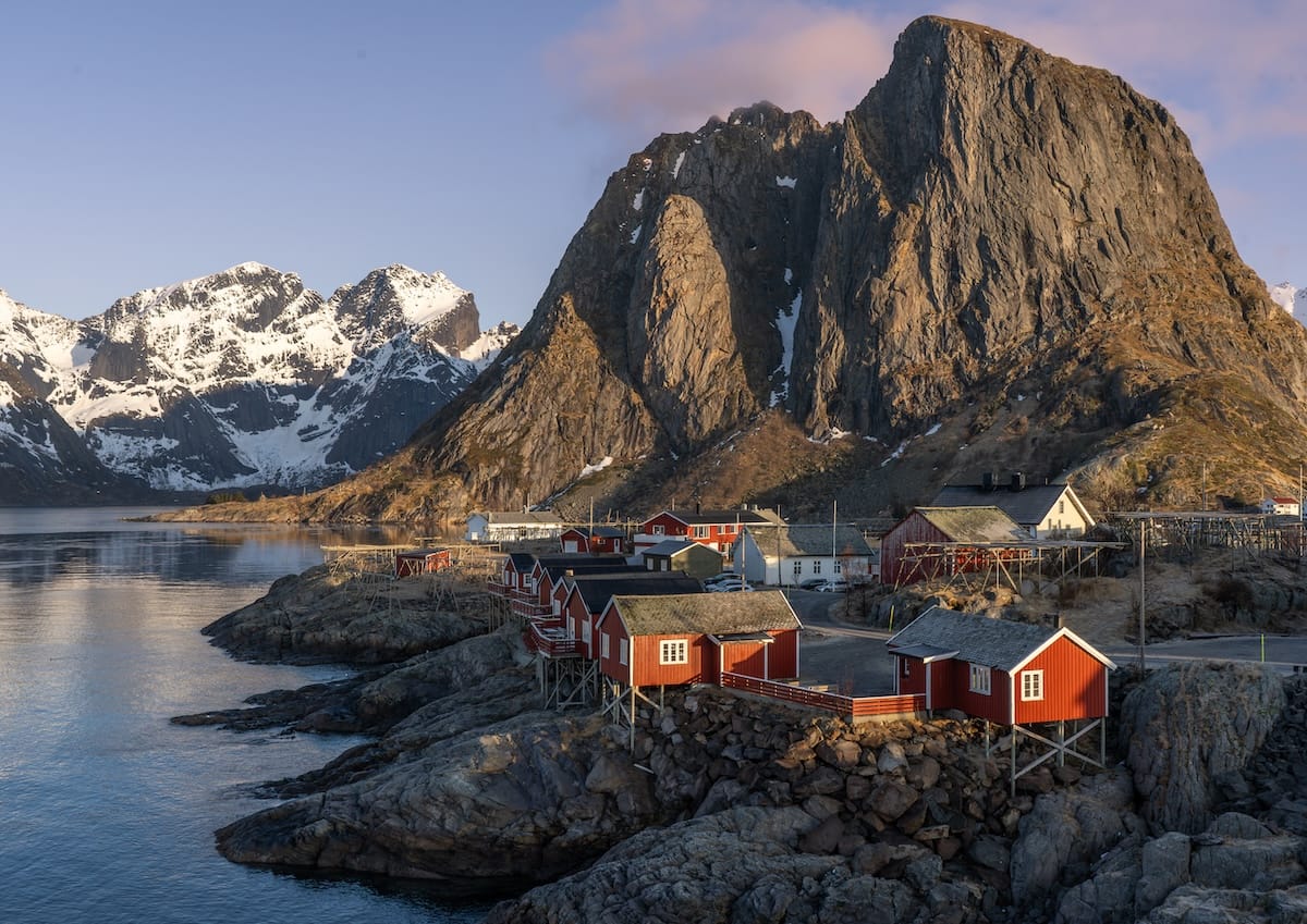 Red rorbu cabins on stilts along the rocky shore of Hamnøy island in Lofoten, beneath the Olstind peak and snowy mountains at sunset