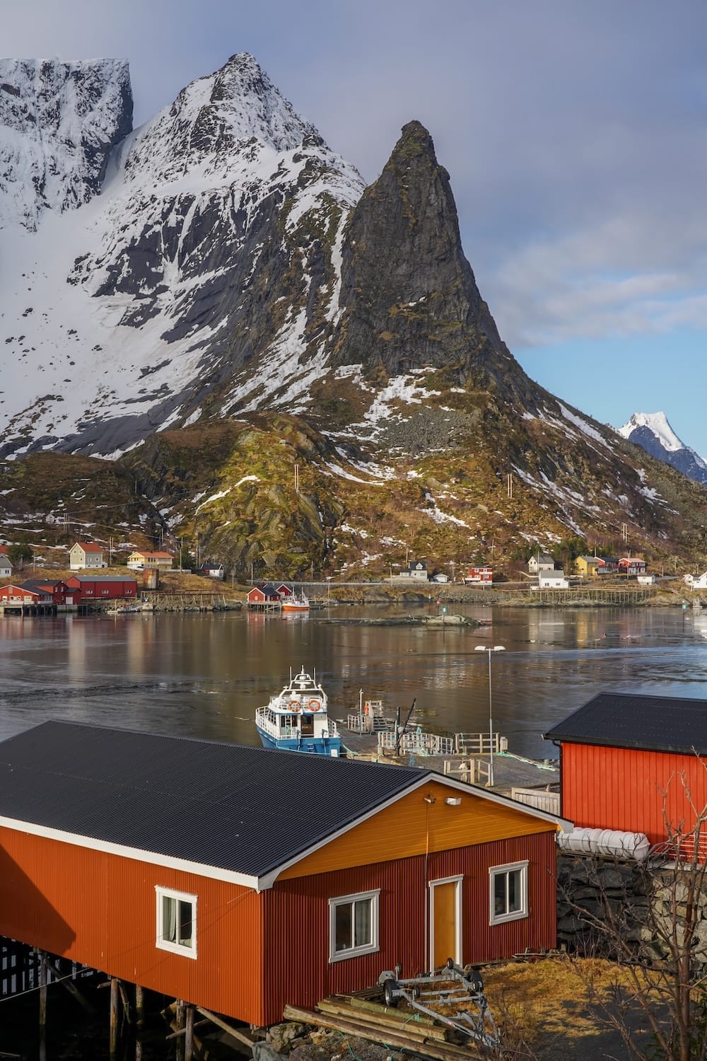 Orange and red rorbu cabins above the harbor in Reine, Lofoten, with a ferry at the dock and a snowy pointed peak behind
