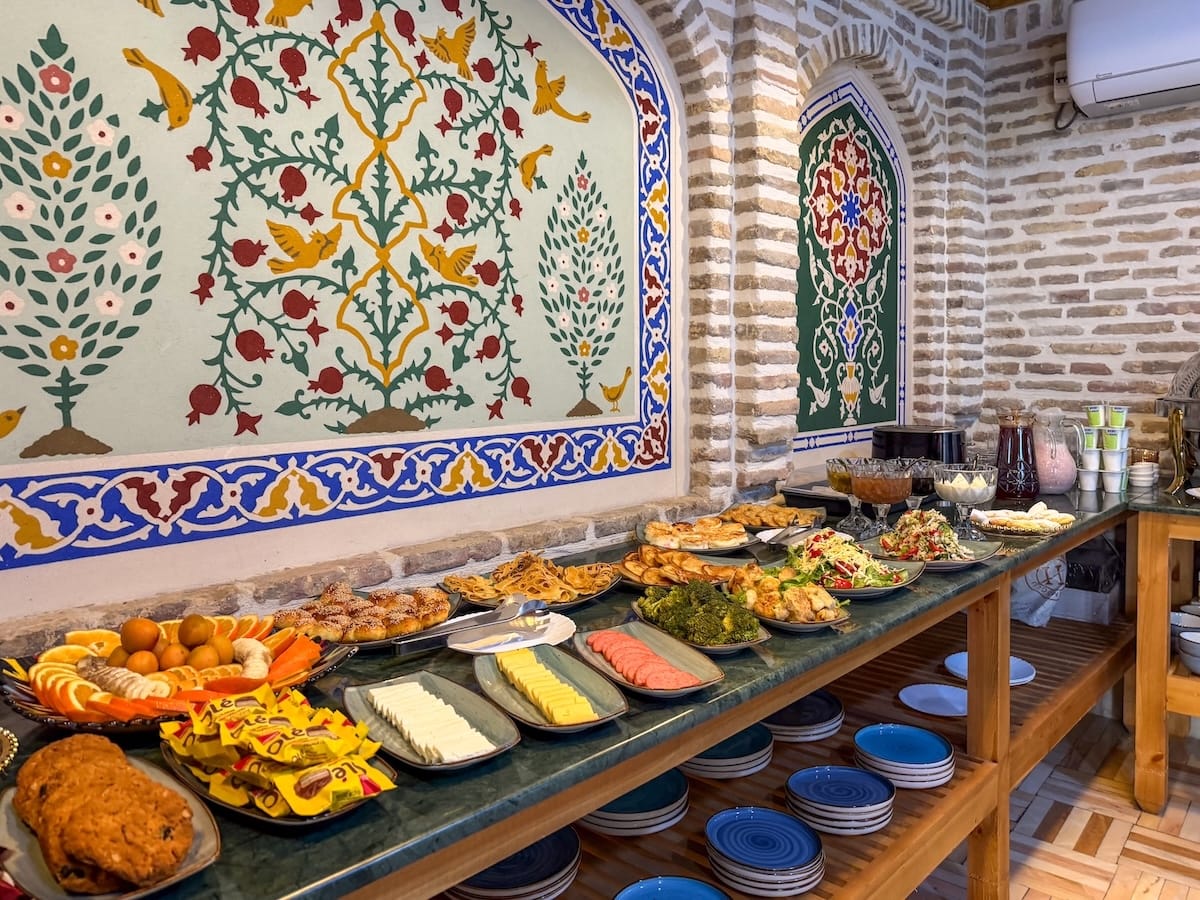 Breakfast buffet with pastries, fruit, and salads beside a painted pomegranate mural at Duston Boutique Hotel in Bukhara, Uzbekistan