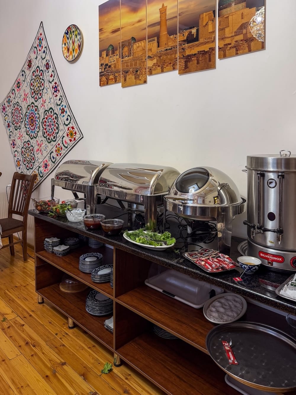 Breakfast buffet with chafing dishes and tea urn beneath a suzani wall hanging at Hotel Rayhon in Bukhara, Uzbekistan