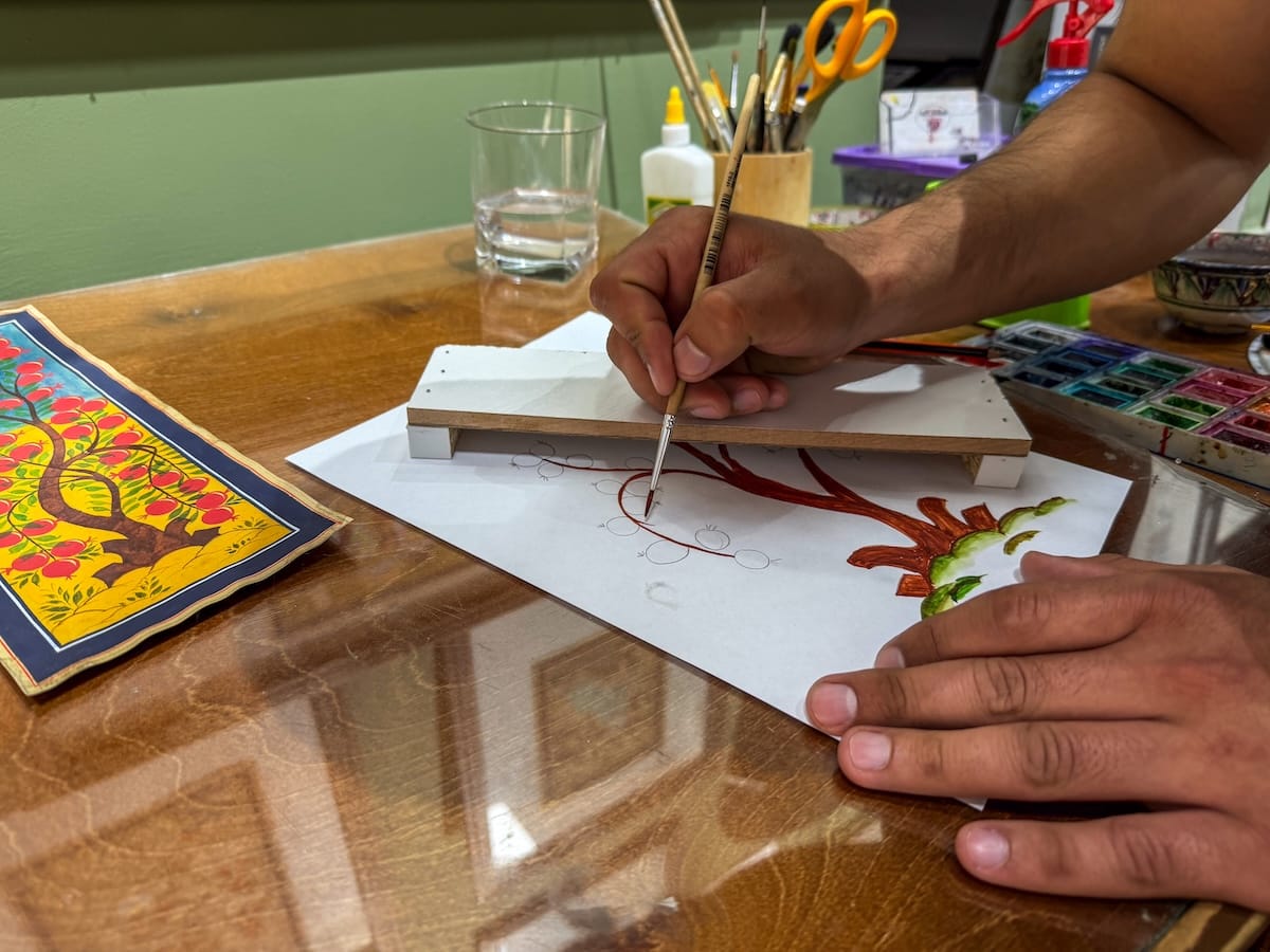 Miniature painter using a fine brush and a hand-rest board to paint a pomegranate tree on paper, with a finished miniature and a watercolor palette on the desk in a Bukhara workshop