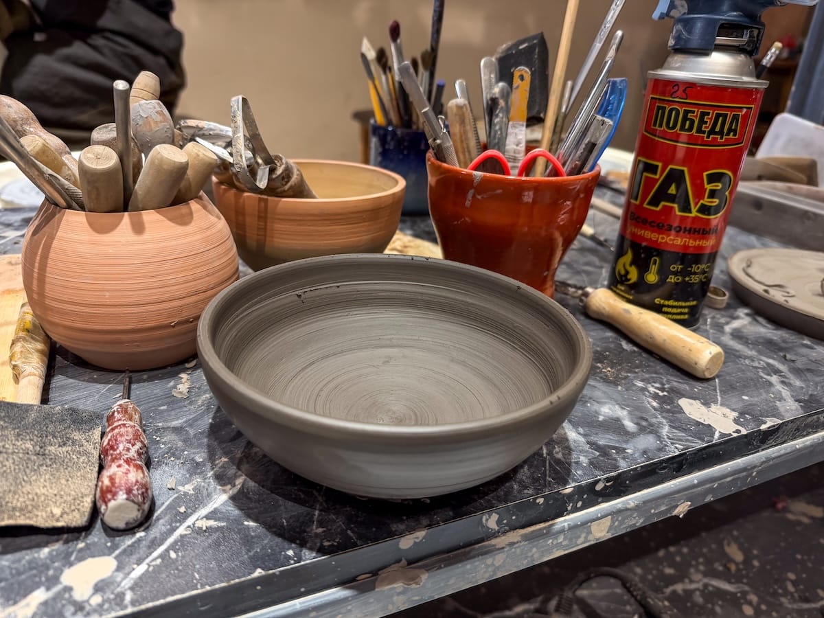 Unfired gray clay bowl on a pottery workbench surrounded by wooden tools and clay pots at a ceramics workshop in Bukhara