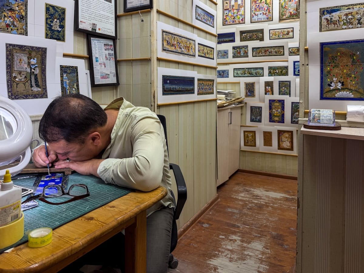Watching Bukharan miniature painter, Firuz, at work
Miniature painter bent close over his desk, painting fine details with a thin brush in a small Bukhara studio, with framed miniature paintings covering the walls behind him