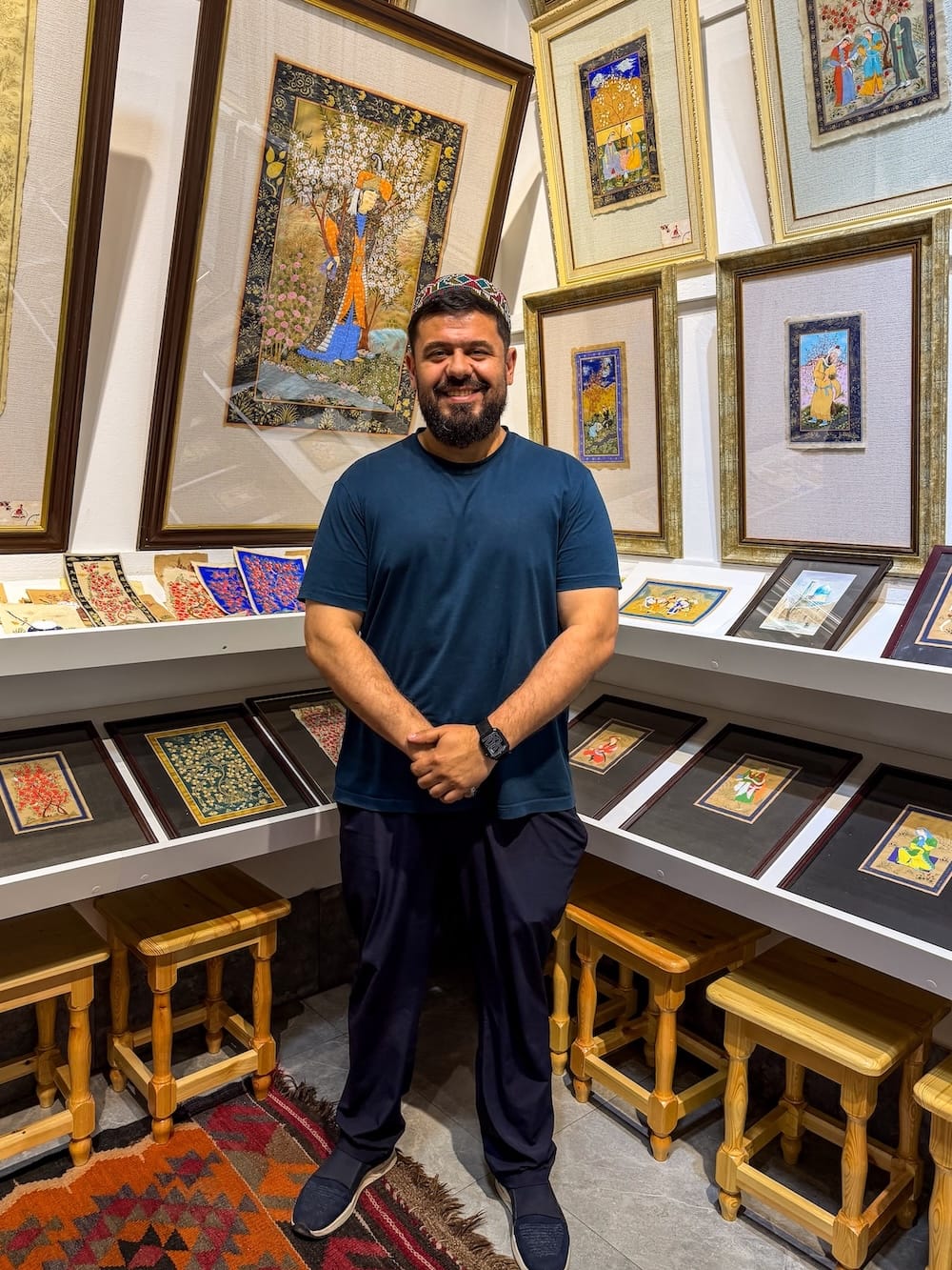 Smiling miniature painting master in a traditional skullcap standing in his Bukhara workshop, surrounded by framed Persian-style miniature paintings and wooden stools