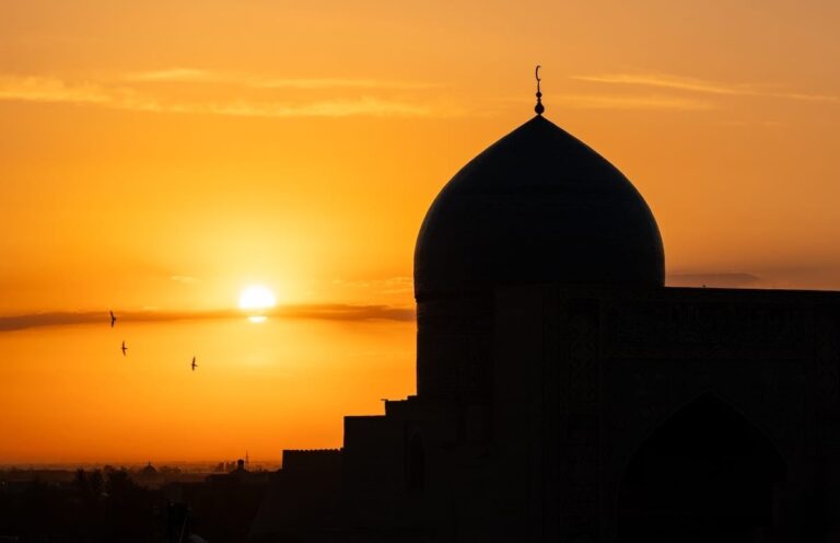 Silhouette of a domed madrasah against an orange sunset with birds flying overhead, Bukhara, Uzbekistan Things to do in Bukhara, Uzbekistan