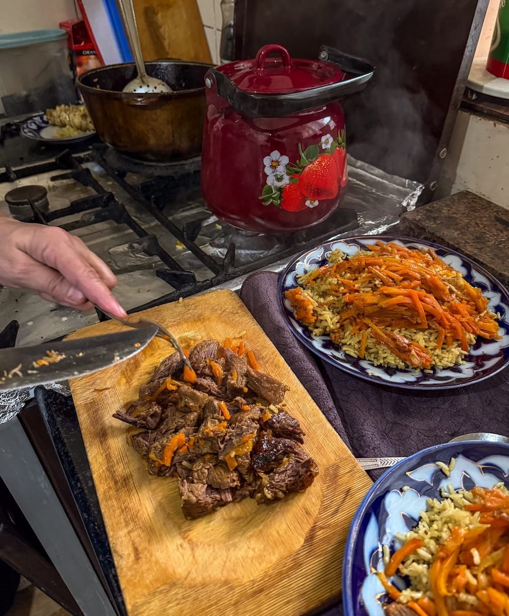 Cooking plov in Bukhara: chopped beef on a wooden board next to plates of rice topped with shredded carrots, served on blue and white ceramic dishes in a home kitchen
