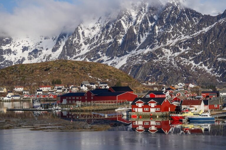 Where to stay in Reine, Sakrisøy, and Hamnøy Red rorbu cabins and boats reflected in calm harbor water below snow-covered mountains in Reine, Lofoten, Norway