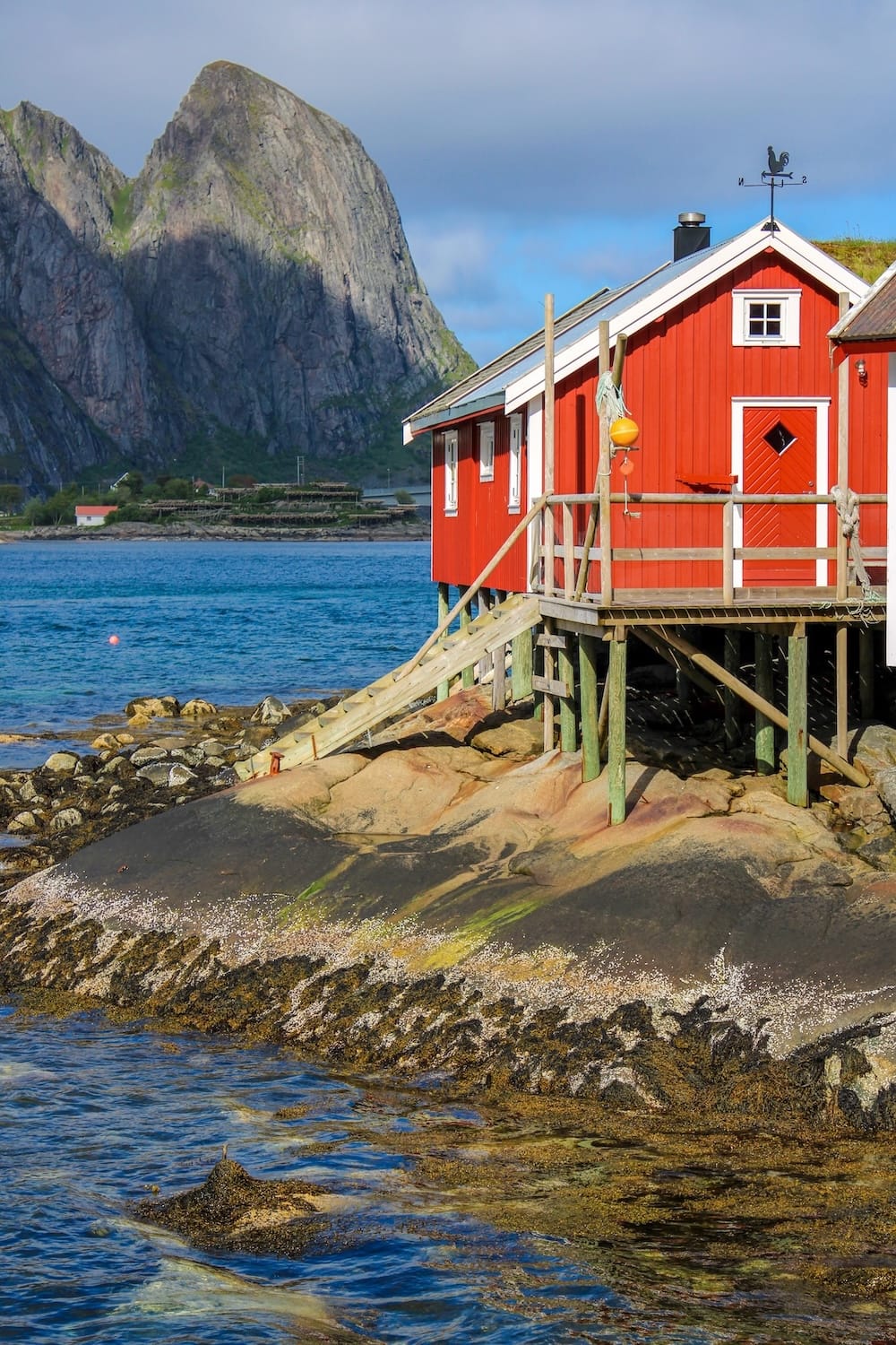 Red rorbu cabin on stilts over rocky shoreline with a steep mountain peak behind it in Reine, Lofoten Islands