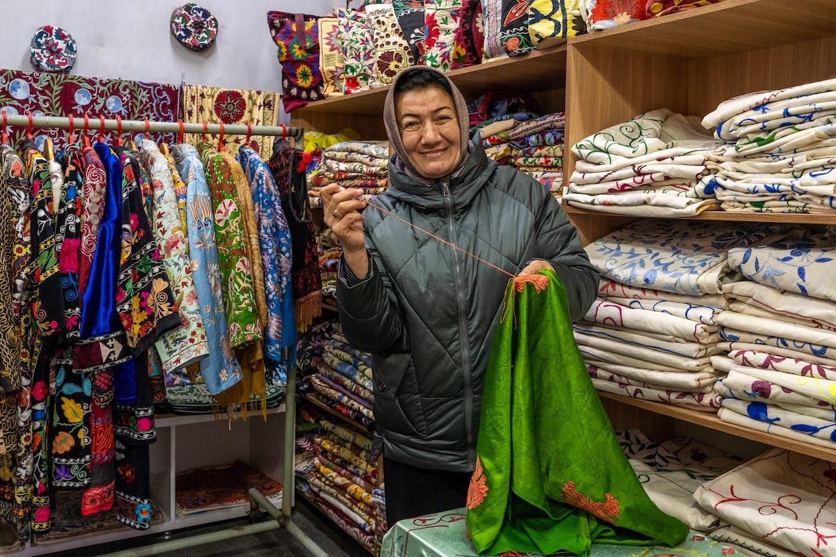 Cultural workshops in Bukhara | Suzani master Nafisa Smiling suzani artisan holding a green silk fabric with orange embroidery in her Bukhara shop, with embroidered coats on a rack and shelves of suzani textiles behind her