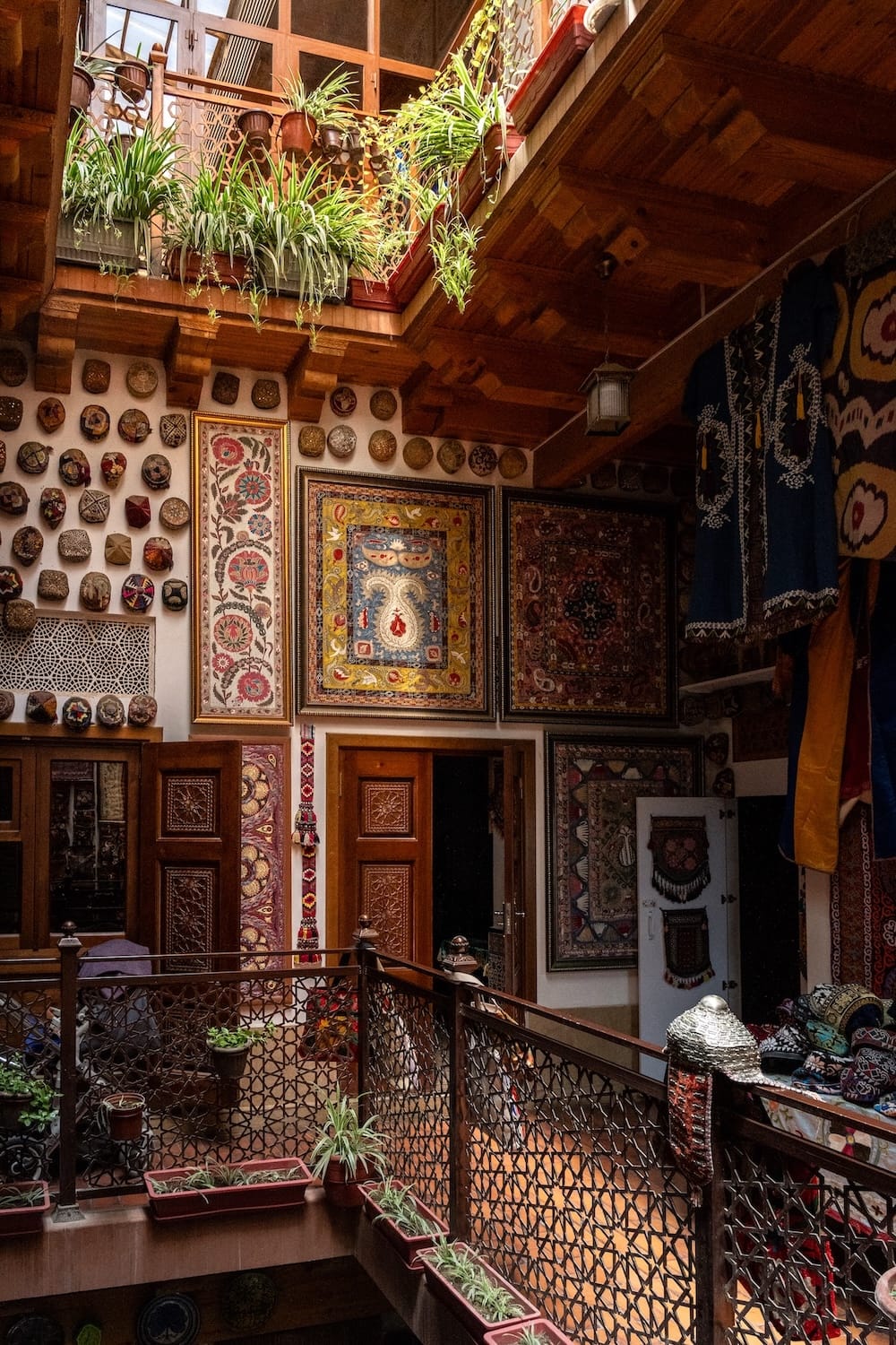 Interior courtyard of a traditional Bukhara craft house with framed suzani embroideries, embroidered skullcaps on the wall, carved wooden doors, and hanging plants on the upper balcony