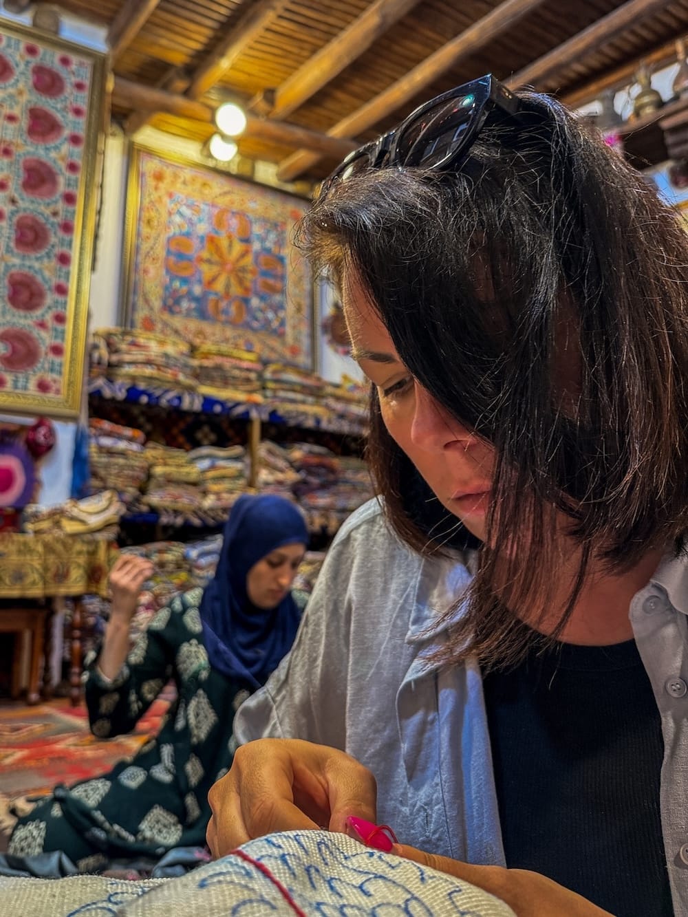 Visitor concentrating on hand-stitching a suzani embroidery design during a workshop in Bukhara, with colorful suzani textiles on the walls and an artisan working in the background