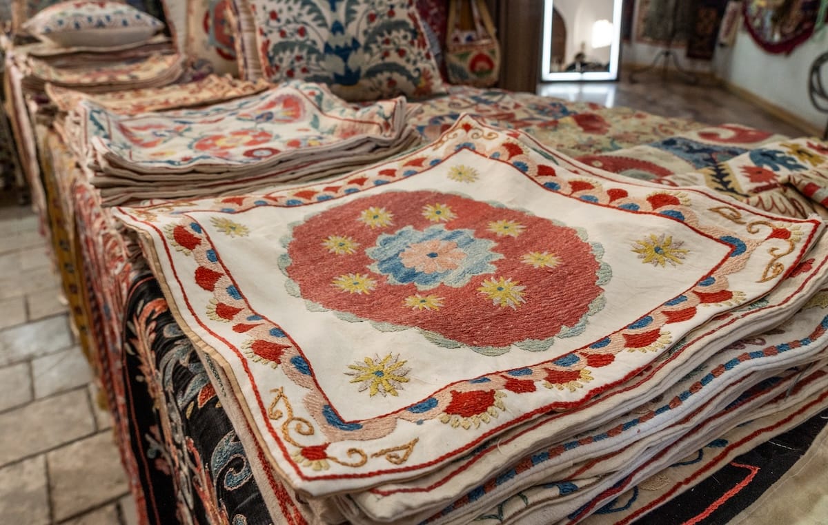 Stacks of handmade suzani embroidered cushion covers in red, blue, and gold floral patterns on display in a Bukhara shop