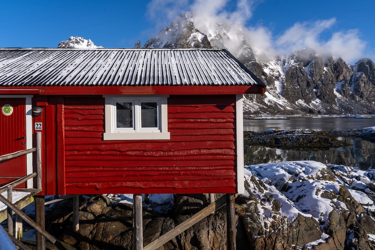 Red rorbu fisherman's cabin on stilts with a snowy roof and mountain behind it in Svolvær