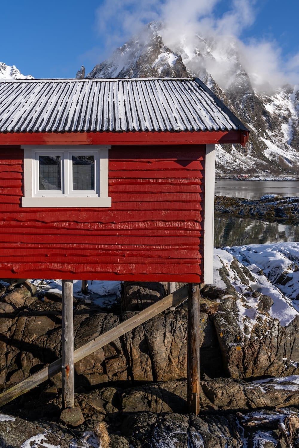 Side view of a red rorbu cabin on stilts over rocks with a cloud-covered mountain peak, Svolvær