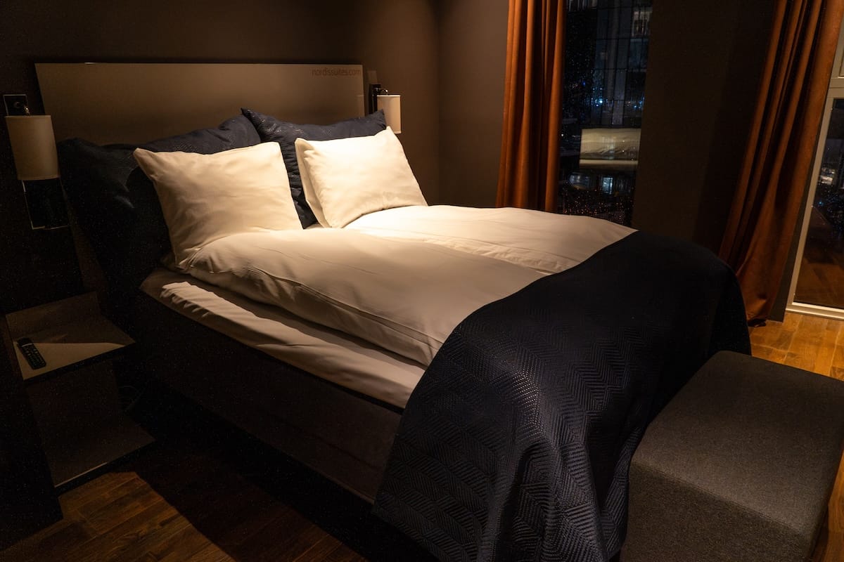 Double bed with white linens and a navy throw in a dark, cozy bedroom at Nordis Apartments, with rust-colored curtains and city lights outside