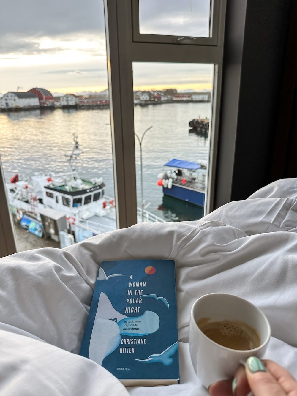 Morning coffee and a book on a bed with a harbor view in Svolvær, Lofoten