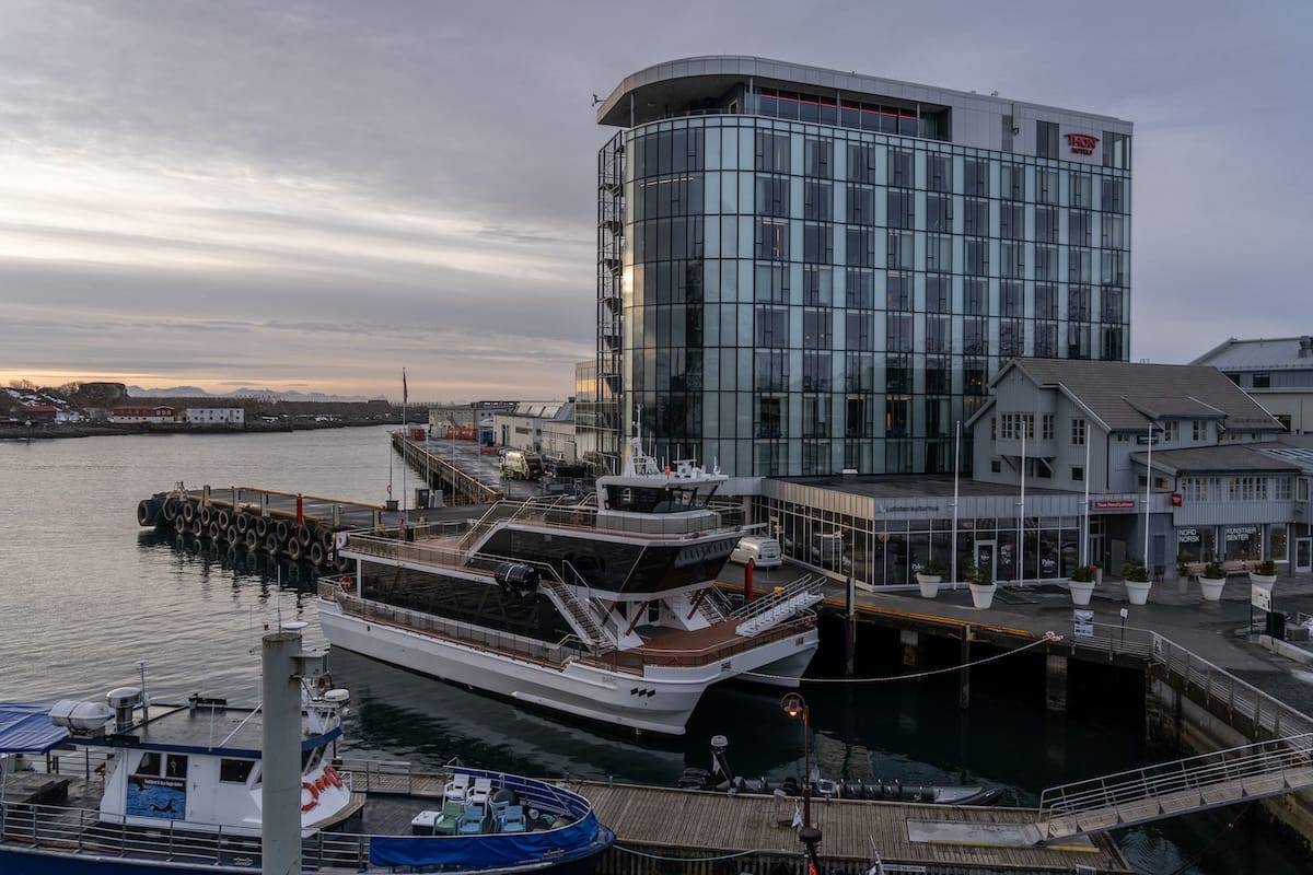 Glass hotel building on the Svolvær waterfront with boats moored in the harbor at sunrise